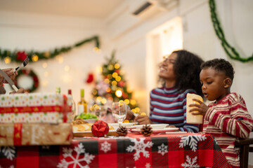 A joyful family gathers around the dinner table celebrating Christmas. The father, dressed in a Santa hat, gives a gift to the child, while everyone smiles with warmth and festive decorations.