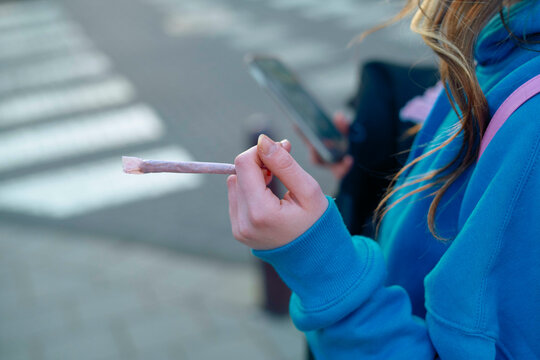 A close-up of a person's hand holding a cigarette or joint while using a smartphone, wearing a blue hoodie.