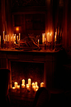 A dimly lit room with multiple candles of varied sizes providing a warm glow, placed on a mantle and the floor.
