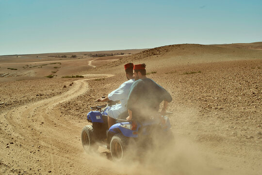 Two individuals ride a blue ATV through a desert landscape, leaving a trail of dust behind them under a clear sky, Morocco