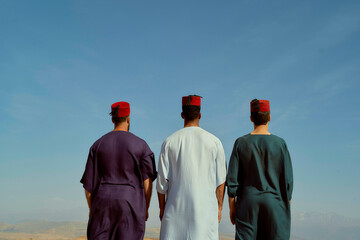 Three men wearing traditional fez hats stand together looking out at a clear sky above a mountainous horizon, Morocco
