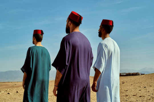 Three men wearing traditional fez hats stand side by side, looking out over a desert landscape, Morocco
