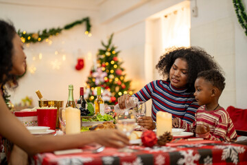 A joyful family gathers around the dinner table celebrating Christmas. The father, dressed in a Santa hat, gives a gift to the child, while everyone smiles with warmth and festive decorations.