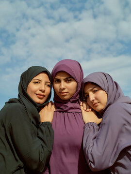 Three women in colorful hijabs standing close together under a blue sky with clouds, Morocco