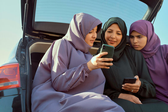 Three women in hijabs looking at a smartphone by a car under a blue sky, Morocco - Powered by Adobe