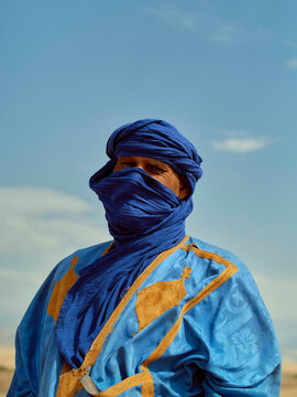 A person wearing a traditional blue turban and adorned in a blue robe with gold accents stands against a clear sky backdrop, Morocco