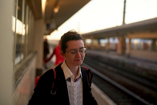 Young woman waiting on a train platform during golden hour, with train tracks and an implied sunset in the background.