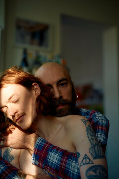 Bald man with beard and shirt embraces a shirtless young man with red hair as sunlight filters through a window, casting shadows on them.