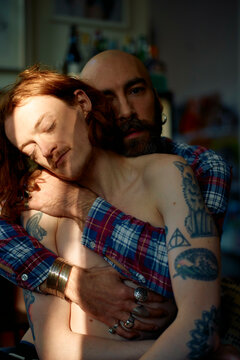 Bald man with beard and shirt embraces a shirtless young man with red hair as sunlight filters through a window, casting shadows on them.