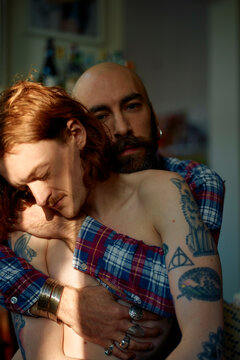 Bald man with beard and shirt embraces a shirtless young man with red hair as sunlight filters through a window, casting shadows on them.
