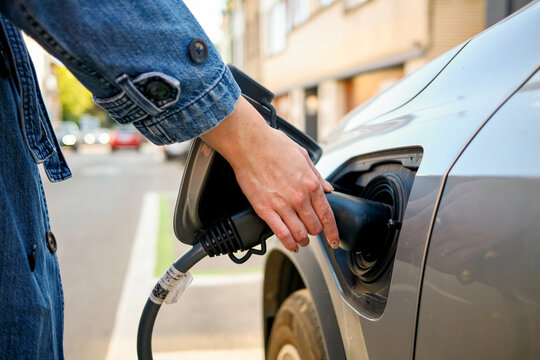Close-up of a person's hand inserting a charging plug into an electric vehicle's charge port on a sunny day.