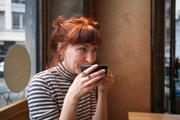 A woman with red hair sipping coffee in a cafe, looking away from the camera with a thoughtful expression, Brussels, Belgium