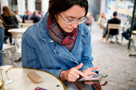 Woman in glasses and denim jacket using smartphone at an outdoor caf� table with a wine glass and wallet beside her.