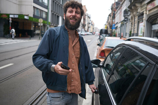 Smiling bearded man in a navy jacket standing next to a car on a city street, holding a smartphone, Brussels, Belgium
