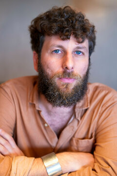Bearded man with curly hair looking at the camera with a neutral expression, wearing a light brown shirt and a silver bracelet.