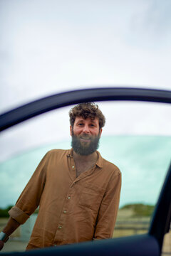 Smiling bearded man standing outside viewed through an open car window with a clear sky in the background.