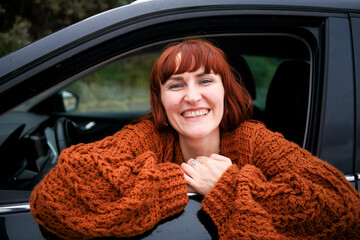Smiling woman with red hair wearing a chunky knit sweater leaning out of a car window.