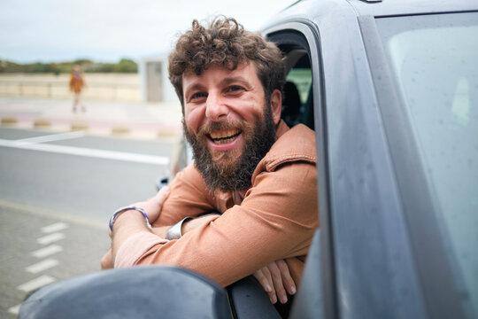 Smiling bearded man leaning out of a car window, exuding a cheerful and relaxed vibe.