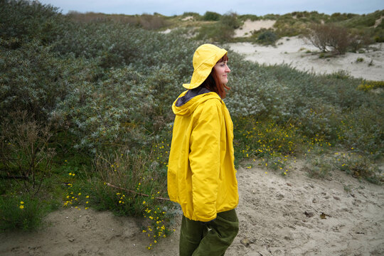 A smiling woman in a yellow raincoat walking through a sandy beach landscape with green shrubbery and yellow flowers, Belgium