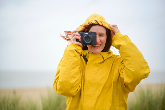 Woman in a yellow raincoat takes a photo with a digital camera on a windy day by the sea, Belgium