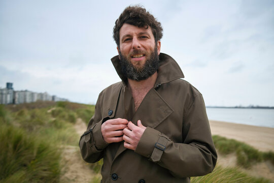 Smiling bearded man buttoning up his coat on a cloudy beach day with city skyline in the background, Belgium