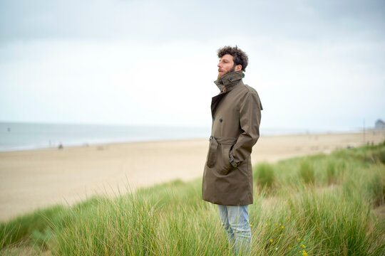 Man in a coat standing in a coastal grassy area with a cloudy sky above and a beach in the background, Belgium