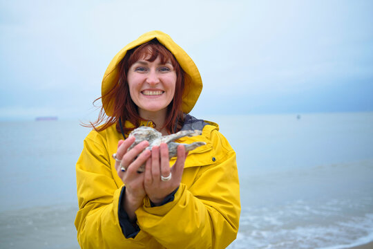 Smiling woman in a yellow raincoat holding an oyster on a foggy beach, Belgium