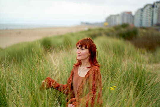 A woman with red hair enjoys a serene moment in a grassy dune on a cloudy day with a beachside cityscape in the background, Belgium