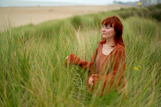 Woman in a brown sweater enjoys the serenity of a grassy dune with a beach and overcast sky in the background, Belgium