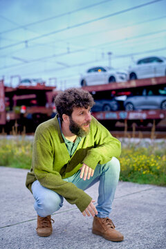 Man in green cardigan and light jeans squatting by a road with a train overhead and cars in the background.
