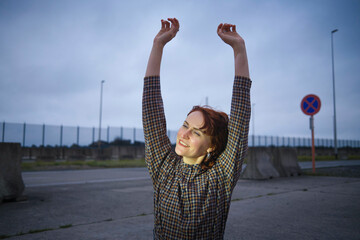 Young woman stretching her arms happily in front of a car at twilight with a no entry sign and an overcast sky in the background.