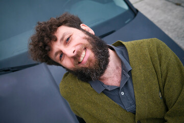 Smiling bearded man in a green jacket leaning casually against a car.