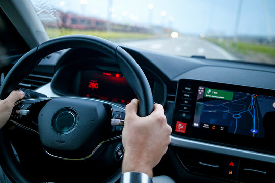Close-up of a driver's hands on a steering wheel with a dashboard and navigation screen in focus.