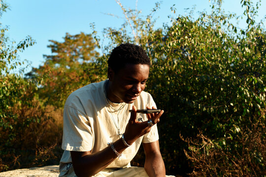 A man sitting outdoors on a rock using a smartphone with trees and blue sky in the background, Berlin, Germany