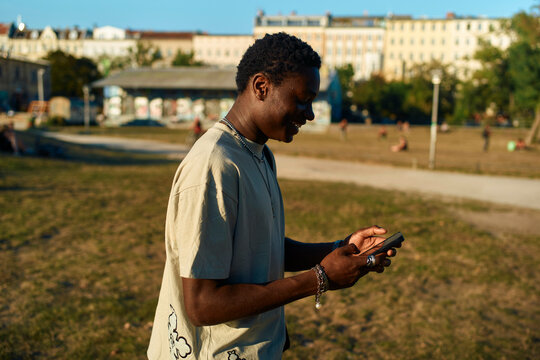 Young man smiling as he uses his smartphone in an urban park at sunset, Berlin, Germany
