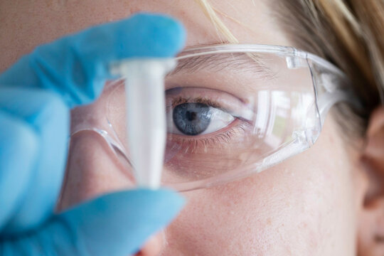 Close-up of a woman's eye looking at a test tube through protective safety glasses in a laboratory setting.