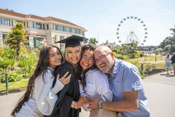 A joyful family group of four celebrates graduation outdoors with a ferris wheel in the background, Bournemouth, Dorset, UK