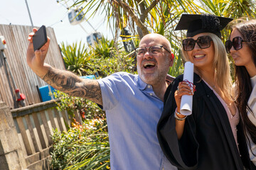 A joyful graduate in a cap and gown takes a selfie with a beaming father and sister under a sunny sky.