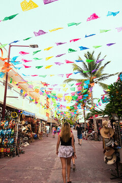 Colorful papel picado banners flutter above a bustling market street with souvenir shops as a woman walks away from the camera.