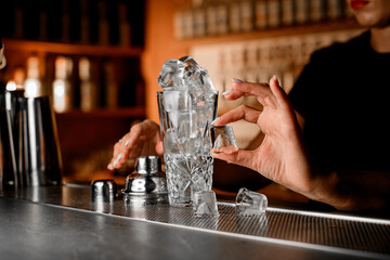 Glass shaker with a slide is filled with ice cubes so that they spill out onto the bar, where they are picked up by female hands