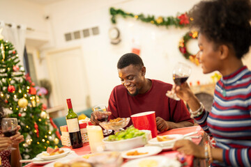 A joyful family shares a Christmas meal together, passing food and creating holiday memories. Decorated tree, festive lights, and presents in the background add warmth to cozy celebration atmosphere.