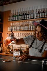 Smiling female bartender pours a cocktail from a mixing glass into another glass, carefully watching the process