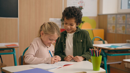 Talking African and Caucasian little school children multiracial diverse girl boy kids pupils schoolchildren together doing task exercise writing in copybook talk help learning class study education