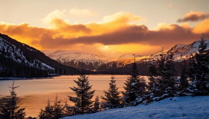 Christmas trees growing behind the mountain lake surrounded by mountains; evening sky and glowing clouds of snow on a hill silhouette with orange sunset light