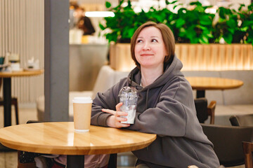 Middleaged woman sitting at caffee in shopping mall and drinking milkshake, while using mobile phone or looking away
