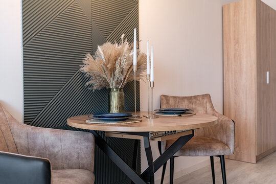 romantic dinner for two by candlelight. There are plates, vase with flowers, candlesticks on round wooden table. Soft brown chairs with back near table. Modern, stylish design of the dining room.