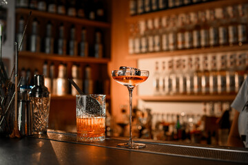 High-stemmed glass filled with a cocktail with a huge ice cube sits on the bar, next to a mixing glass