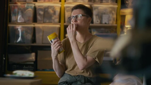 Medium slowmo shot of female worker in glasses eating sandwich and drinking hot tea from thermo mug during lunch break in garage workshop
