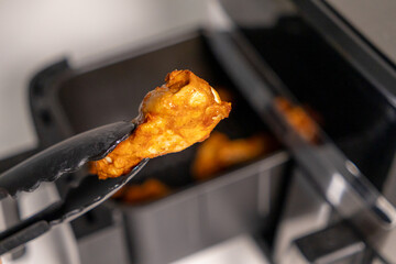 Close-up of a crispy chicken wing held with tongs above air fryer, perfectly cooked and golden