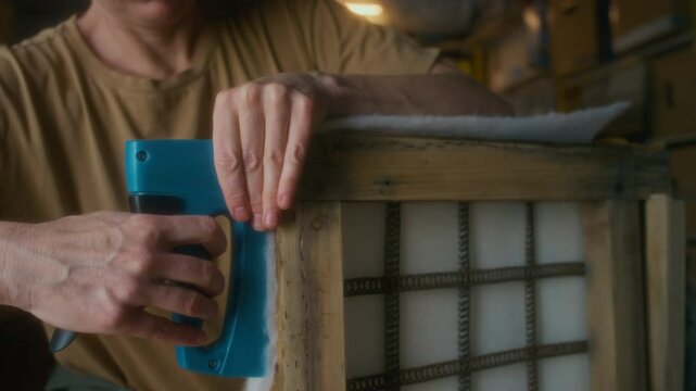 Closeup of hands of female upholsterer stapling soft wadding to wooden chair framework using construction stapler while reupholstering and restoring furniture in workshop